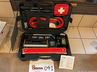 Front view of the open black hard plastic storage case showing all items inside: jumper cables, compressor, tools, first aid kit, and reflective triangle.