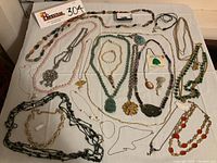 Full view of assorted necklaces, bracelets, pins, laid out on white cloth showing variety of pieces including green stone carved pendants, pearls, chains, and bead necklaces.