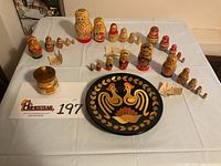 All items arranged on white cloth showing the five sets of nesting dolls, rooster plate, and village scene pot with lid. Receipts visible near dolls.