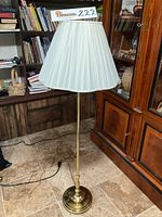 Full view of the floor lamp showing pleated cream fabric shade, brass pole with trumpet motif near base, and circular brass base.