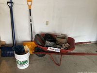 Wide shot of wheelbarrow, shovels, broom, and bucket with various items inside wheelbarrow placed against wall.