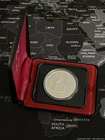 Silver coin in black capsule seated in red velvet-lined display case with hinged lid, showing coin details and packaging.