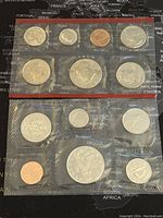 Two proof-like US and Canadian coin sets in sealed plastic holders on a world map background, showing all coins in each set clearly.