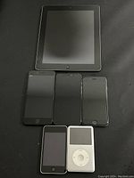 Full view of all six devices including iPad, three iPhones, iPod Touch, and iPod Classic on black surface