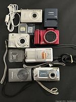 Top-down view of all digital cameras and accessories laid out on black background showing various brands and models along with cords and memory cards.