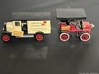 Two die cast model vehicles side by side on black surface: a vintage farm produce delivery truck and early Cadillac automobile model.