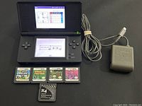 Nintendo DS console open with screens powered on showing clock/calendar and Pictocraft interface with power cord connected, placed on black surface alongside 5 game cartridges and R4 cartridge.