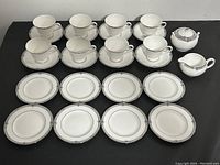 Wide view of all items: 8 teacups on saucers, 8 dessert plates laid out, creamer, and sugar bowl with lid all with white bone china, blue floral and geometric border, silver trim