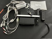 Photo of Nintendo Wii console with power adapter, sensor bar, wired black Wii Remote with wrist strap, AV cables, and operation manual laid out on table.