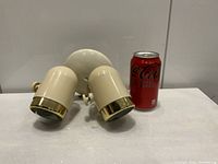 Front view of vintage 1970s swivel spotlight wall lamp with two cream colored cylindrical heads and gold-toned trim, positioned sideways with a soda can for size reference.