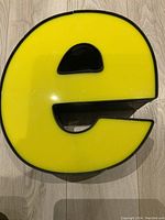 Front view of the yellow and black vintage acrylic letter 'e' placed flat on a light wood floor, showing its shape and color.