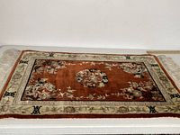 Asian silk rug laid flat showing rust red central field with floral and bird motifs, cream and green decorative borders with black symbols.