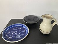 Three piece set of pottery with a blue fish plate, dark bowl, and cream pitcher on black background.