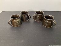 Four Ian Steele pottery mugs with brown and green glaze, arranged showing shape and handles.