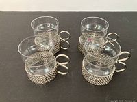 Four clear Iittala Tsaikka glasses with textured metal holders and looped handles shown from front angle on black surface