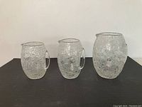 Front view of three crackle glass pitchers in small, medium, and large sizes displayed side by side on a flat surface.