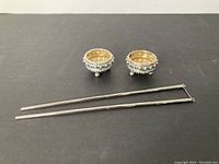 Two small 800 silver dishes with beaded decoration and gold-toned interior next to a pair of silver chopsticks connected by a chain, on a black surface.
