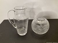Photo of the clear Versace by Rosenthal crystal pitcher with etched Greek key pattern and Waterford round crystal vase with diamond cut pattern on black surface against white background.