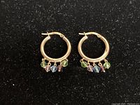 Frontal view of both gold hoops with five colored gemstone dangles each against black background