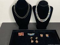 Photo showing two beaded necklaces displayed on black velvet bust stands with five pairs of earrings and brooches arranged below.