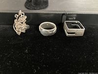 Three silver rings placed side by side on black velvet background. Includes floral, plain band and square-shaped designs.