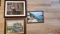 Photo showing all three framed original artwork pieces arranged on floor: bird painting top left, railway house painting bottom left, coastal sea pathway painting right
