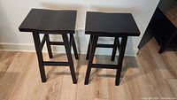 Two dark brown wooden bar stools standing on a light wooden floor against a white wall.