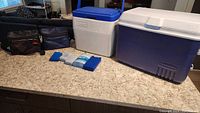 Closed view of a large cooler, small cooler, two lunch boxes in black cases, and ice packs set on a kitchen counter.