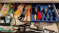 Photo of kitchen drawer containing assorted cutlery and utensils including forks, spoons, knives, scissors, tongs, peelers, and other tools.
