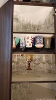 Front view of cabinet shelf showing multiple clear glass beer and wine glasses, several ceramic mugs with colorful patterns and plain finishes.