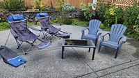 Outdoor setting showing two navy blue folding camp chairs with footrests and carry cases alongside two grey Adirondack plastic chairs and a rectangular glass top coffee table.