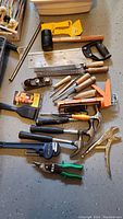 Photo of assorted hand tools including saws, hammers, chisels, pliers, caulking gun and others arranged on gray surface.