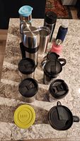 Multiple travel mugs and water bottles arranged on a granite counter, showing variety in size, color, and lid design.