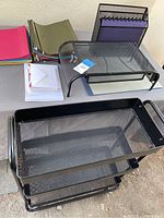 Photo showing a black mesh metal desk riser, a black folder holder filled with purple hanging folders, assorted colored folders in red, pink, green, and some clear sleeves on the table next to the art cart.