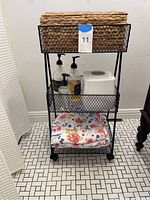 Black metal three-tier cart with woven baskets on all tiers, holding two soap/lotion dispensers, white tissue box on middle tier, and floral cloth on bottom tier.