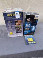 Photo of two bug zapper boxes and one fruit fly trap on gray cloth background.