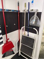 Photo of multiple cleaning tools hanging on wall including brooms with dustpans and brushes along with a white 2-step foot stool folded closed