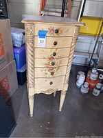 Front view of tall wooden jewelry cabinet with floral motif, carved columns, multiple drawers, and tapered legs.