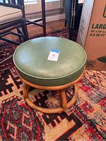 Full view of the rattan footstool with green vinyl cushion on patterned rug near chairs and cardboard box.