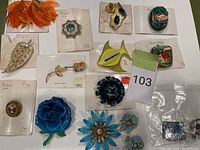 Photo of assorted vintage clip-on earrings and brooches including floral and bird motifs mostly on display cards, laid out on white surface.