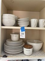 Photo showing six white bowls, six smaller plates, six larger plates, three white mugs, a white creamer labeled 'CREAM', and a white sugar bowl with wooden lid labeled 'SUGAR' on shelf.