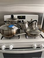 Photo showing assortment of pots and pans including pans with glass lids and handles, arranged on a white stove.