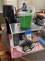 SodaStream machine, salad spinner, napkin holder, and box of wine glasses on table.