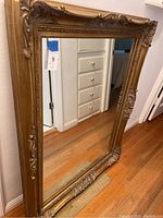 Full view of the large vintage mirror leaning against wall showing overall size and ornate gold frame with carved details.