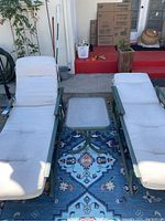 Two green metal framed chaise lounges with light beige cushioned seats and backs arranged side by side with a square frosted glass top side table between them positioned on a dark patterned outdoor rug. Outdoor cushions show signs of wear and need cleaning.