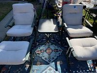 Front view of two green metal outdoor chairs with white cushions, two matching ottomans with white cushions, and a rectangular table with dark textured top placed on a patterned outdoor rug.