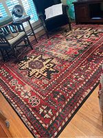 Full view of the rug laid out in a living room showing intricate geometric and floral tribal patterns in red, black, and cream.