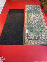 Photo showing both rugs side by side on red surface. Black runner is plain and ribbed, green runner has floral pattern.
