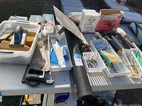 Wide view of the tools and household items on a table outdoors, showing multiple containers and loose tools including a hand saw, putty knives, sponge and a large black tube.