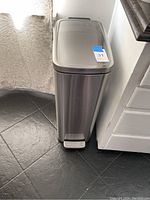 Front view of tall stainless steel step trash can positioned against a wall near a kitchen counter.